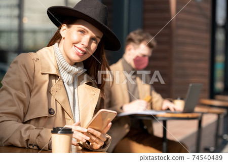 Young brunette businesswoman in hat, sweater and trenchcoat using smartphone Young brunette businesswoman in hat, sweater and trenchcoat using smartphone 74540489