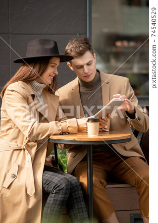 Young restful couple sitting outdoors, having drinks and discussing online news Young restful couple sitting outdoors, having drinks and discussing online news 74540529