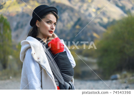 Beautiful young mother with her infant baby in sling outdoor. Mother is carrying her child and travel in autumn mountain. Babywearing concept Beautiful young mother with her infant baby in sling outdoor. Mother is carrying her child and travel in autumn mountain. Babywearing concept 74543398