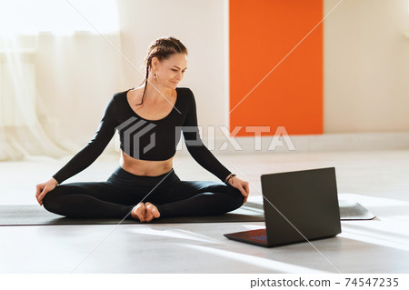 A woman in black sportswear practicing yoga, looking at a laptop, performs the Baddha Konasana exercise, butterfly pose. Concept on the topic of online training A woman in black sportswear practicing yoga, looking at a laptop, performs the Baddha Konasana exercise, butterfly pose. Concept on the topic of online training 74547235