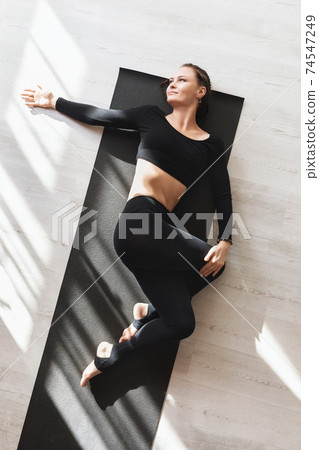A woman in black sportswear practicing yoga while lying on a gymnastic mat performs the Supta Matsyendrasana exercise with twisting her legs into the garudasana position 74547249