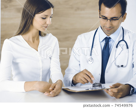 Doctor and patient discussing medical exam results while sitting at the desk in clinic. Male physician using clipboard for filling up medication history record of young woman. Data in medicine 74549976