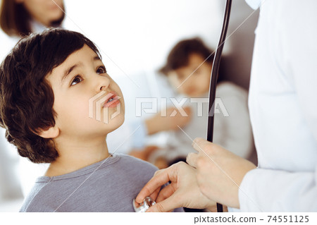 Doctor-woman examining a child patient by stethoscope. Cute arab boy and his brother at physician appointment. Medicine concept 74551125