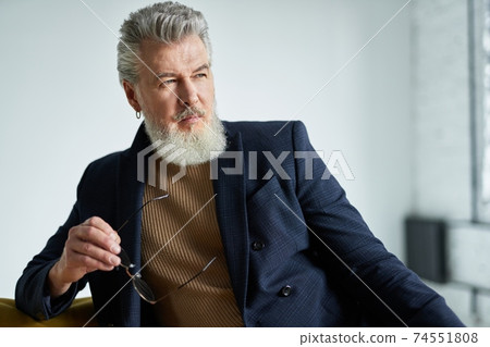 Portrait of stylish serious middle aged man with beard looking away, holding glasses while sitting on sofa indoors 74551808