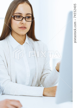Young woman sitting at the desk with computer in white colored office. Looks like student girl or business lady communicating with casual dressed man Young woman sitting at the desk with computer in white colored office. Looks like student girl or business lady communicating with casual dressed man 74552405