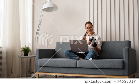 Happy young woman with photo camera using laptop at home Happy young woman with photo camera using laptop at home 74553317