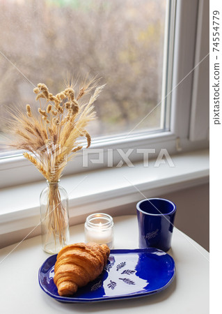 Handmade blue clay plate with croissant on it. Vase with wheat and candles. Handmade blue clay plate with croissant on it. Vase with wheat and candles. 74554779