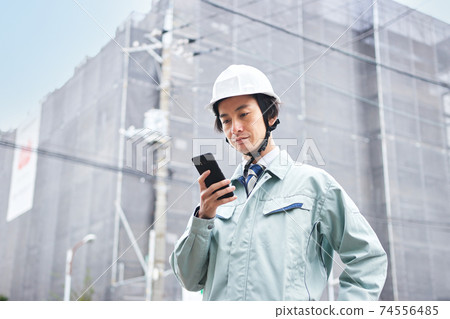 一個穿著工作服的男人看著一個建築工地前的智能手機 74556485
