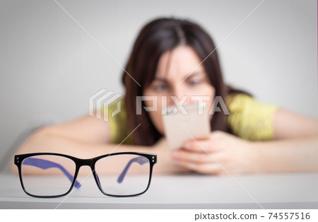In the foreground there are glasses for vision, in the background a girl squints into a smartphone. The concept of harm to eyes and vision, health In the foreground there are glasses for vision, in the background a girl squints into a smartphone. The concept of harm to eyes and vision, health 74557516