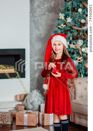 One cute cheerful girl santa hat near Christmas tree. Holds red christmas ball. 74560661