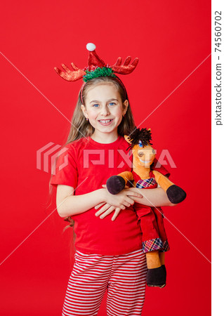 Portrait of a funny cheerful girl with a bandage of horns on her head hugging a teddy horse in Christmas pajamas isolated on a bright red background. 74560702