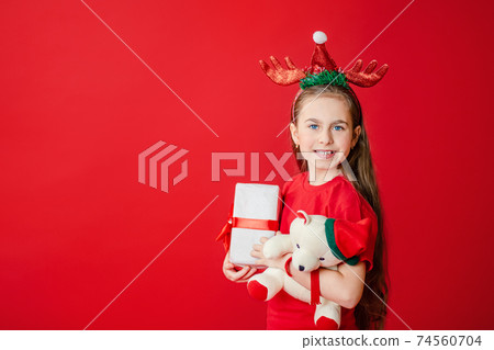 Portrait of a funny cheerful girl with a bandage of horns on her head hugging a teddy bear in Christmas pajamas isolated on a bright red background. 74560704