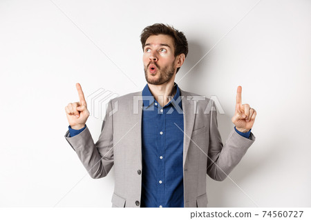 Amazed bearded guy in business suit pointing and looking up wondered, say wow and showing exciting thing, standing against white background 74560727