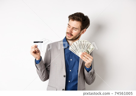 Successful man holding dollar bills and looking at plastic credit card, pleased with bank, standing on white background in business suit Successful man holding dollar bills and looking at plastic credit card, pleased with bank, standing on white background in business suit 74561005