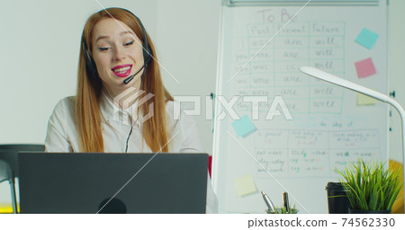 Woman in headphones talking to students via video conference at empty class. Smiling female school teacher having online class indoors. Education concept. Quarantine concept. Study by internet. 74562330