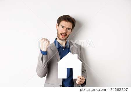 Real estate. Excited man buying house and showing paper home cutout, say yes and fist pump with joy, standing in suit on white background Real estate. Excited man buying house and showing paper home cutout, say yes and fist pump with joy, standing in suit on white background 74562797