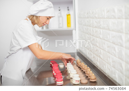 cooking, food and baking concept. Pretty woman confectionery chef making colorful tasty macarons, connect the halves with ganache cream at modern kitchen in pastry shop 74563084