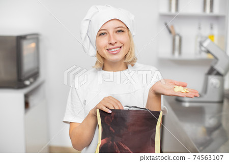 Young smiling Caucasian woman pastry chef, holding bag with white chocolate drops and demonstrating drops in hand to camera. Making of desserts Young smiling Caucasian woman pastry chef, holding bag with white chocolate drops and demonstrating drops in hand to camera. Making of desserts 74563107