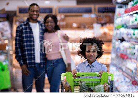 Happy little black girl with shopping trolley and her parents buying goods at modern supermarket, free space Happy little black girl with shopping trolley and her parents buying goods at modern supermarket, free space 74564018
