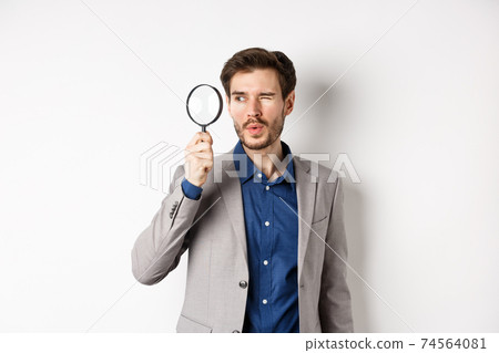 Handsome male model in suit looking through magnifying glass with interest, seeing something aside, white background Handsome male model in suit looking through magnifying glass with interest, seeing something aside, white background 74564081