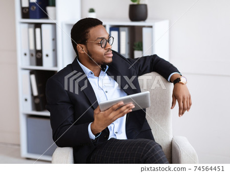 Thoughtful black businessman sitting in chair, using digital tablet Thoughtful black businessman sitting in chair, using digital tablet 74564451