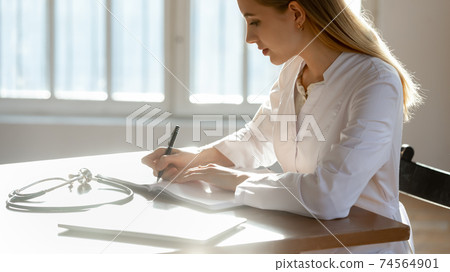 Concentrated young female doctor making records in paper copybook. 74564901