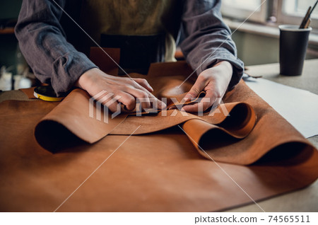 A young apprentice in a boot workshop prepares leather for further use on a large table 74565511