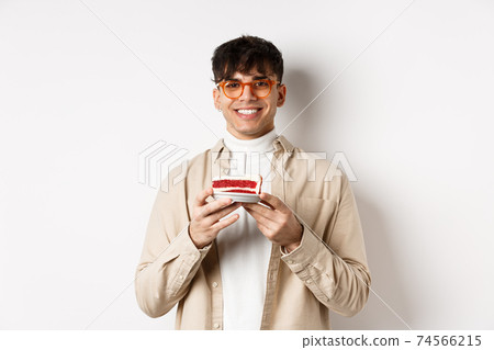 Real people and holidays concept. Happy hipster guy in glasses celebrating birthday, making wish on b-day cake with candle and smiling, standing on white background Real people and holidays concept. Happy hipster guy in glasses celebrating birthday, making wish on b-day cake with candle and smiling, standing on white background 74566215