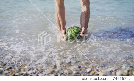 A young guy stands by the sea and holds a ripe watermelon in his hands. The guy gets a watermelon out of the sea. A young guy stands by the sea and holds a ripe watermelon in his hands. The guy gets a watermelon out of the sea. 74566762