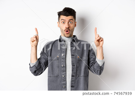 Excited man checking out promo offer, gasping amazed and pointing fingers up, making cool announcement, standing on white background 74567939