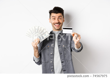 Image of handsome guy holding dollar bills but suggesting use plastic credit card, smiling friendly at camera, standing on white background 74567975