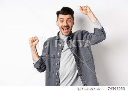 Excited man shouting from joy, winning prize and rejoicing, raising hands up and yelling yes with satisfied face, triumphing, achieve goal, standing on white background 74568953