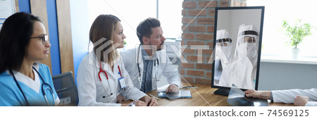 Doctors at meeting communicating via video communication with colleagues in protective overalls Doctors at meeting communicating via video communication with colleagues in protective overalls 74569125