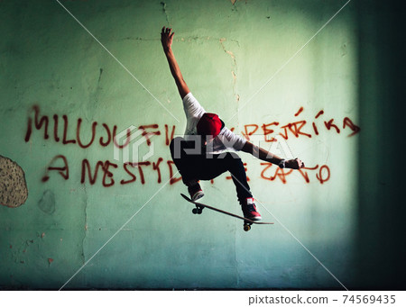 Young skateboarder caught in peak moment of his stunt with arms outstretched, board lightly touching shoes. In background wall of room in abandoned house with sprayed text. Natural light conditions. 74569435