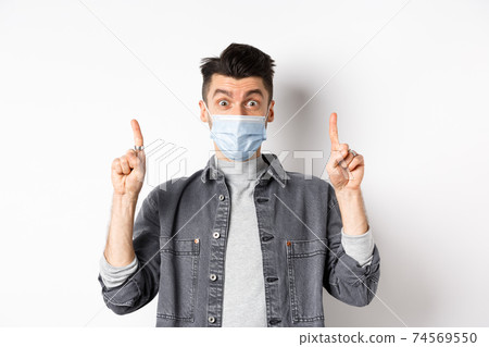 Pandemic lifestyle, healthcare and medicine concept. Excited man advertising in medical mask, pointing fingers up and look amazed, standing on white background 74569550