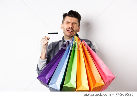 Sad crying man wasted all money on shopping, holding bags and showing empty credit card, standing against white background 74569585