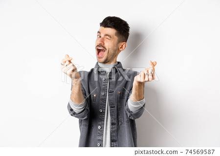Funny guy winking and showing hand hearts sign, feeling romantic and happy, standing on white background Funny guy winking and showing hand hearts sign, feeling romantic and happy, standing on white background 74569587