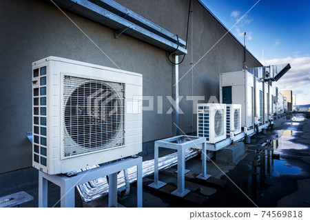 Square air-conditioning unit on the roof with a round fan. In the background gradually receding other units that are out of focus. On the right side light blue sky and commercial space. Square air-conditioning unit on the roof with a round fan. In the background gradually receding other units that are out of focus. On the right side light blue sky and commercial space. 74569818