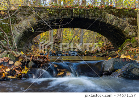 A Water cascade in the creek flowing under an ancient stone bridge in the autumn forest. A Water cascade in the creek flowing under an ancient stone bridge in the autumn forest. 74571157