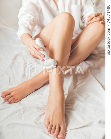 Woman takes care of herself, depilated legs with epilator. Epilation cosmetological procedure for hair removal. Spa at home. Skin care. Woman takes care of herself, depilated legs with epilator. Epilation cosmetological procedure for hair removal. Spa at home. Skin care. 74571486