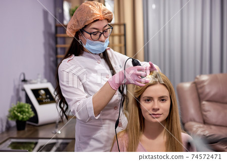 Trichological examination. young woman with thinning hair at examination of the scalp and hair with a microscope Trichological examination. young woman with thinning hair at examination of the scalp and hair with a microscope 74572471