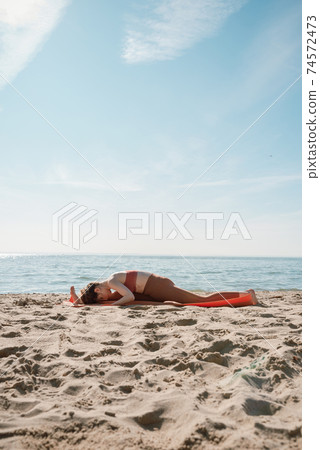 Young female in orange top and leggins practising Yoga split variation (Hanumanasana) on a beach in the morning Young female in orange top and leggins practising Yoga split variation (Hanumanasana) on a beach in the morning 74572473