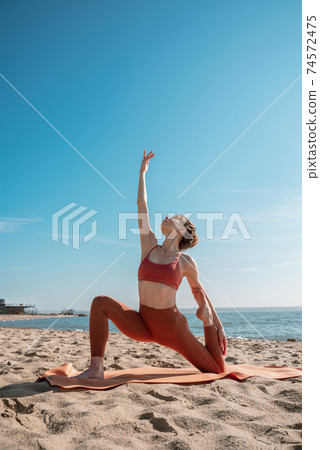 Young female in orange top and leggins practising Crescent Moon Pose (Anjaneyasana Variation) on a beach in the morning Young female in orange top and leggins practising Crescent Moon Pose (Anjaneyasana Variation) on a beach in the morning 74572475