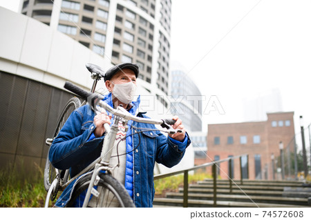 Senior man carrying bicycle outdoors on street in city, coronavirus concept. Senior man carrying bicycle outdoors on street in city, coronavirus concept. 74572608