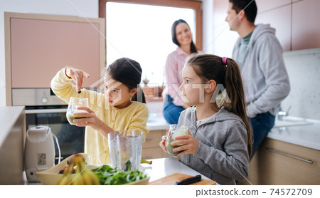 Small girls with parents indoors at home, preparing fruit smoothie drink. Small girls with parents indoors at home, preparing fruit smoothie drink. 74572709