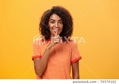 Not say anyone till I give permission. Happy pleased and mysterious attractive dark-skinned female with curly hairstyle telling beauty secret to friend making shh sound with index finger over mouth 74574102