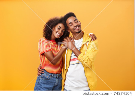 They love each other. Portrait of two charming african american man and woman in relationship hugging with heartwarming smile touching hands smiling gently posing against orange background 74574104