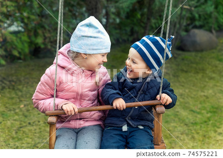 Two cute adorable playful caucasian siblings boy girl child enjoy having fun swinging wooden swing at backyard together with father. Little toddler brother and sister laughing at playing outdoors 74575221