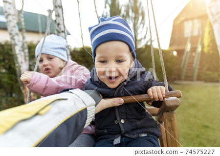 Two cute adorable playful caucasian siblings boy girl child enjoy having fun swinging wooden swing at backyard together with father. Little toddler brother and sister laughing at playing outdoors 74575227
