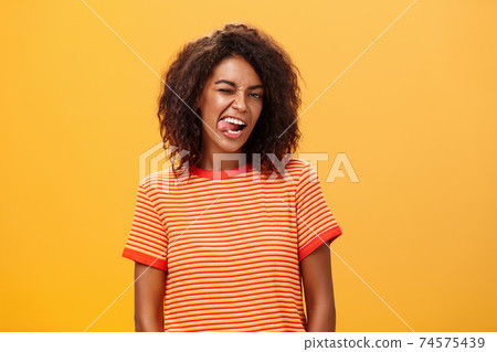 Portrait of daring and emotive confident flirty woman with afro hairstyle winking joyfully showing tongue posing carefree and enthusiastic against orange background flirting with hot guy 74575439
